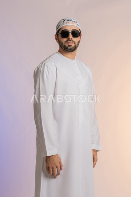 الأناقة والوسامة والاهتمام بالمظهر الخارجي، بورتريه لرجل عربي خليجي اماراتي  يرتدي الكندورة والطاقية يقف  بإيماءات الثقة بالنفس ويضع نظارة شمسية سوداء، استخدام العدسات الطبية لحماية العين من أشعة الشمس، خلفية ملونة