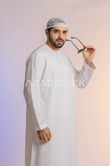 الأناقة والوسامة والاهتمام بالمظهر الخارجي، بورتريه لرجل عربي خليجي اماراتي يرتدي الكندورة والطاقية يقف بإيماءات الثقة بالنفس ويضع نظارة شمسية سوداء، استخدام العدسات الطبية لحماية العين من أشعة الشمس، خلفية ملونة