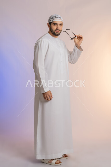 الأناقة والوسامة والاهتمام بالمظهر الخارجي، بورتريه لرجل عربي خليجي اماراتي يرتدي الكندورة والطاقية يقف بإيماءات الثقة بالنفس ويضع نظارة شمسية سوداء، استخدام العدسات الطبية لحماية العين من أشعة الشمس، خلفية ملونة