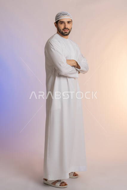 الاهتمام بالأناقة والمظهر الخارجي، النظر إلى الكاميرا بايماءات الجدية والسرور، مفهوم الرجولة والقوة والثقة بالنفس، بورتريه لرجل عربي خليجي اماراتي يرتدي الكندورة  والقبعة يقف بأيدي متقاطعة بتعابير السعادة والسرور، خلفية ملونة