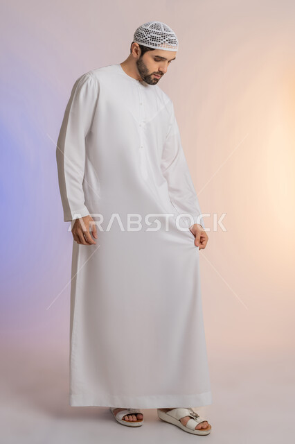 الاهتمام بالمظهر الخارجي, مفهوم الأناقة والرجولة, بورتريه لرجل عربي خليجي اماراتي يرتدي الكندورة ويضع القبعة يمسك ثوبه بيده، الوقوف باستقامة واعتدال، خلفية ملونة