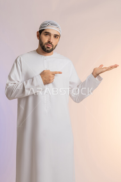 رفع وحمل عنصر معين، تحديد شيء معين بإيماءات السرور والسعادة، بورتريه لرجل عربي خليجي اماراتي يرتدي الكندورة والقبعة البيضاء يفتح كفه للأعلى ويشير إلى عنصر ما, حمل عنصر هولوجرامي، خلفيه ملونة