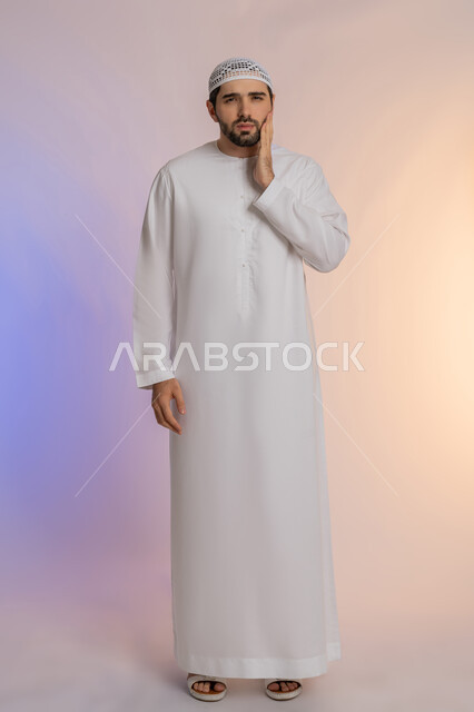 مشاكل التهاب اللثة وتسوس الأسنان, الاشارة إلى الفم بتعابير الإرهاق والتعب, بورتريه لرجل عربي خليجي اماراتي يرتدي الكندورة والقبعة ينظر الى الكاميرة بإيماءات الانزعاج والألم الشديد، صورة الجسم بالطول الكامل، خلفية ملونة
