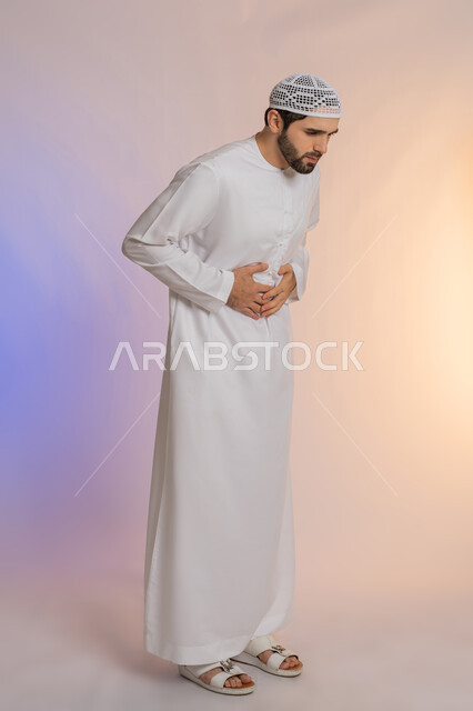 Indigestion and a feeling of heartburn, the need for medication and a treatment plan, a feeling of cramping, fatigue and exhaustion, a portrait of an Arab Gulf Emirati man wearing a kandora and a hat holding his stomach with pain gestures, full-length body portrait, colored background.
