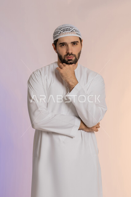 Looking forward and planning for the future, solving problems and making decisions, portrait of an Emirati Gulf Arab man wearing a kandura and a hat, placing his hand on his chin with gestures of thought and absent-mindedness, the concept of listening and focusing, a colorful background.