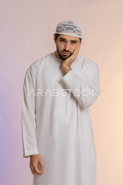 Pointing to the mouth with expressions of exhaustion and fatigue, problems with gingivitis and tooth decay, a portrait of an Arab Gulf Emirati man wearing a kandora and a hat looking at the camera with gestures of discomfort and severe pain, a colored background.