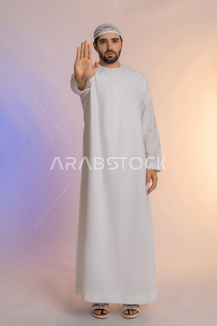 A warning against doing something, a request to stop and not approach, the concept of refusal, abstention and discomfort, a portrait of an Emirati Gulf Arab man wearing a kandura and a hat, extending his palm forward with gestures prohibiting doing something, full-length body, colored background.