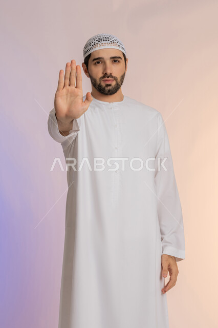The concept of rejection, abstention, and discomfort, a warning against doing something, a request to stop and not approach, a portrait of an Arab Gulf Emirati man wearing a kandura and a hat, extending his palm forward with gestures prohibiting doing something, colored background.