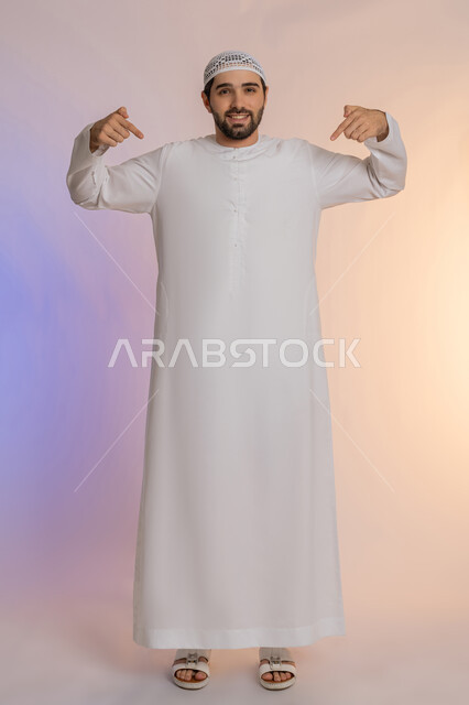 تأكيد حدوث شيء هذه اللحظة, حركات تدل على هذا المكان (هنا)، تحديد موقع معين، بورتريه لرجل عربي خليجي اماراتي يرتدي الكندورة والقبعة يشير بإصابع السبابة نحو الأسفل, النظر إلى الكاميرا بايماءات السرور، الطول الكامل للجسم, خلفية ملونة