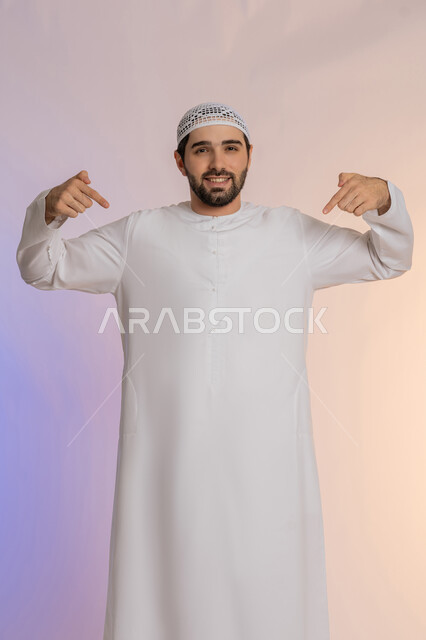 تحديد موقع معين، تأكيد حدوث شيء هذه اللحظة, حركات تدل على هذا المكان (هنا)، بورتريه لرجل عربي خليجي اماراتي يرتدي الكندورة والقبعة يشير بإصابع السبابة نحو الأسفل, النظر إلى الكاميرا بايماءات السرور, خلفية ملونة