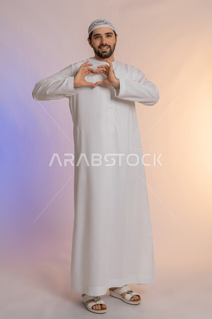 التعبير عن المحبة والمودة، إظهار الحب للآخرين، الاهتمام بالمظهر الخارجي، مفهوم الأناقة والوسامة، النظر إلى الكاميرا بإيماءات البشاشة، بورتريه لرجل عربي خليجي اماراتي  يرتدي الكندورة والقبعة يقوم بصنع شكل قلب بيديه، الجسم بالطول الكامل، خلفية ملونة