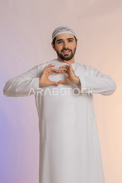 إظهار الحب للآخرين، التعبير عن المحبة والمودة، الاهتمام بالمظهر الخارجي، مفهوم الأناقة والوسامة، النظر إلى الكاميرا بإيماءات البشاشة، بورتريه لرجل عربي خليجي اماراتي  يرتدي الكندورة والقبعة يقوم بصنع شكل قلب بيديه، خلفية ملونة