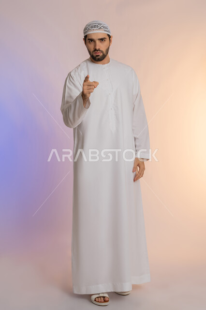 الإشارة باصبع السبابة إلى الكاميرا بشكل مباشر، التهديد والتوعد بتنفيذ شيء ما، إيماءات الترهيب والتحذير، توجيه أصابع الاتهام إلى شخص معين، بورتريه لرجل عربي خليجي اماراتي يرتدي الكندورة والقبعة يشير إلى مكان ما محدد، الجسم بالطول الكامل، خلفية ملونة