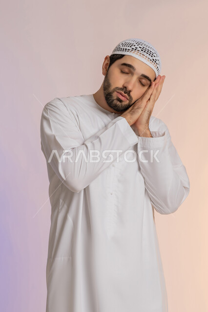 Feeling sleepy and needing to sleep, closing the eyes and placing the hands under the cheeks, the need to take some rest and relaxation, a portrait of an Arab Gulf Emirati man wearing a kandora and a hat, closing his eyes with gestures of fatigue and exhaustion, a colored background.