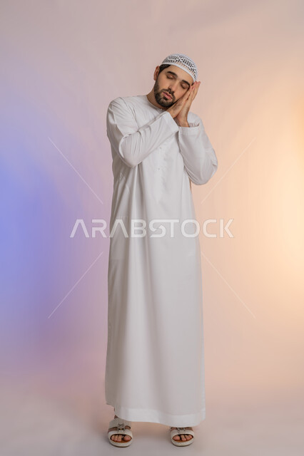 إغماض العينين ووضع اليدين أسفل الخد، الشعور بالنعاس والحاجة للنوم، ضرورة أخذ قسط من الراحة والاسترخاء، بورتريه لرجل عربي خليجي اماراتي يرتدي الكندورة والقبعة يغلق عينيه بإيماءات التعب والإرهاق، الجسم بالطول الكامل، خلفية ملونة