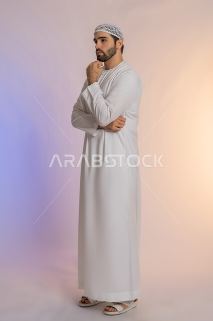Thinking deeply and absent-mindedness, trying to find a solution to difficult problems, anxiety and confusion in making the right decision, portrait of an Emirati Gulf Arab man wearing a kandora and a hat, placing his hand under his chin and looking somewhere with gestures of integration, body full length, colored background.