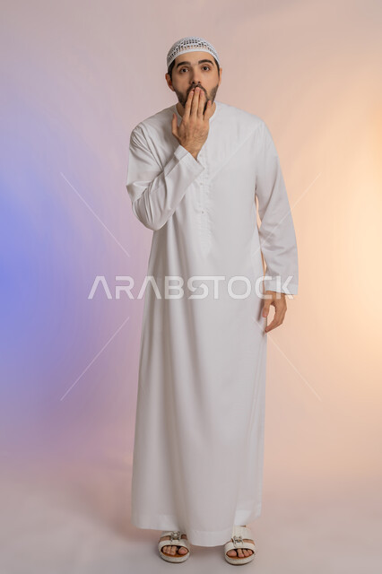 Gestures of astonishment and surprise, exposure to shock and disbelief, concern for outward appearance, disappointment and negative feelings, portrait of an Emirati Gulf Arab man wearing a kandura and a hat, placing his hand in front of his mouth, full-length body, colored background.