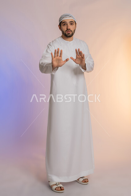 طلب التوقف عن فعل شيء ما، الأمر بالوقوف وعدم التمادي، الرفض وعدم الخضوع، النهي عن التقدم وأهمية ترك مسافة أمان، بورتريه لرجل عربي خليجي اماراتي يرتدي الكندورة والقبعة البيضاء يرفع كفه للأعلى بإيماءات الغضب والجدية،  خلفية ملونة