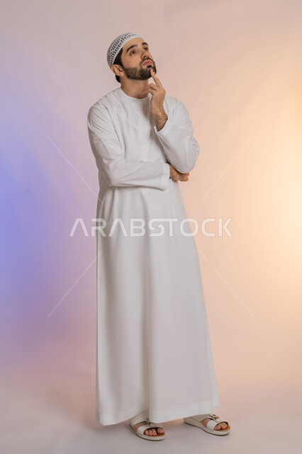 Looking forward and planning for the future, solving problems and making decisions, portrait of an Arab Gulf Emirati man wearing a kandura and a white hat, putting his hand on his chin with gestures of thinking and absent-mindedness, the concept of listening and concentration, colored background