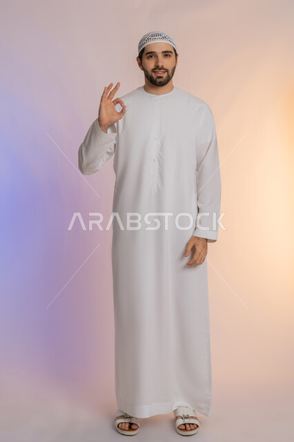 رفع اليد بعلامات الجودة والقبول، التعبير عن الإعجاب والتقدير، الامتياز والعلامة الكاملة، الشعور بالرضا والاستحسان، بورتريه لرجل عربي خليجي اماراتي يرتدي الكندورة والقبعة البيضاء ينظر إلى الكاميرا بإيماءات السعادة والسرور، الجسم بالطول الكامل، خلفية ملونة