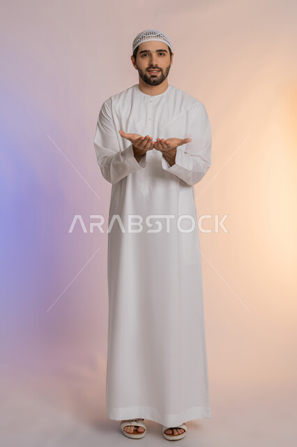 فتح الكفين ومدهما محو الأمام، تقديم الضيافة أو إعطاء شيء ما، الأناقة والوسامة والرجولة، مفهوم التقدير والاحترام، بورتريه لرجل عربي خليجي اماراتي يرتدي الكندورة والقبعة البيضاء ينظر إلى الكاميرا بإيماءات السعادة والسرور،  خلفية ملونة