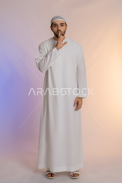 طلب التوقف عن الكلام والصمت، النهي والزجر والانصات، وضع اصبع السبابة أمام الفم بإيماءات الجدية، الأمر بالصمت التام،  بورتريه لرجل عربي خليجي اماراتي يرتدي الكندورة والقبعة البيضاء ينظر للكاميرا بإيماءات الغضب، الجسم بالطول الكامل، خلفية ملونة