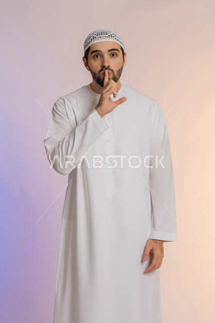 وضع اصبع السبابة أمام الفم بإيماءات الجدية، طلب التوقف عن الكلام والصمت، النهي والزجر والانصات، الأمر بالصمت التام،  بورتريه لرجل عربي خليجي اماراتي يرتدي الكندورة والقبعة البيضاء ينظر للكاميرا بإيماءات الغضب، خلفية ملونة