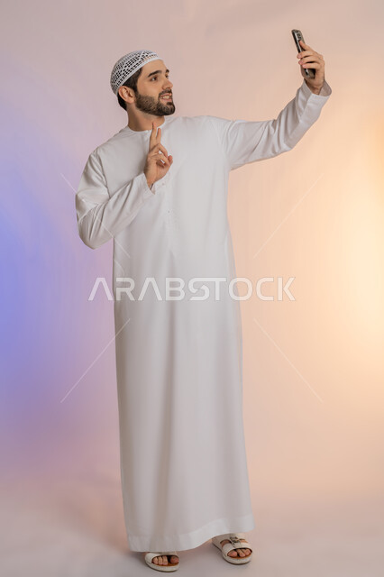 رفع علامة النصر والسلام، اجراء مكالمة فيديو والتواصل مع الاهل، التقاط صورة سيلفي تذكارية باستخدام الهاتف المحمول، إشارة الفوز والانتصار، تحقيق الأهداف والطموحات، بورتريه لرجل عربي خليجي اماراتي يرتدي الكندورة والقبعة يمسك الجوال، الجسم بالطول الكامل، خلفية ملونة