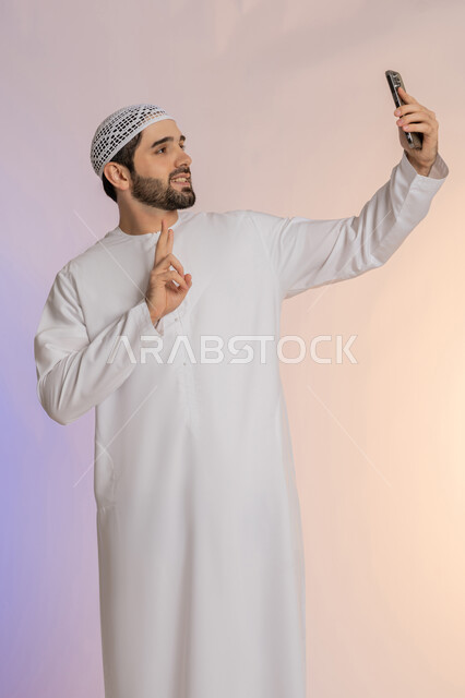 تحقيق الأهداف والطموحات، رفع علامة النصر والسلام، اجراء مكالمة فيديو والتواصل مع الاهل، التقاط صورة سيلفي تذكارية باستخدام الهاتف المحمول، إشارة الفوز والانتصار، بورتريه لرجل عربي خليجي اماراتي يرتدي الكندورة والقبعة يمسك الجوال، خلفية ملونة