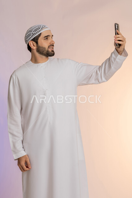 اجراء مكالمة فيديو والتواصل مع الاهل والاصدقاء، التقاط صور السيلفي باستخدام الهاتف المحمول، دمج التقنيات والتكنولوجيا الحديثة بالحياة اليومية، أخذ اللقطات الشخصية التذكارية، بورتريه لرجل عربي خليجي اماراتي يرتدي الكندورة والقبعة يمسك الجوال، خلفية ملونة