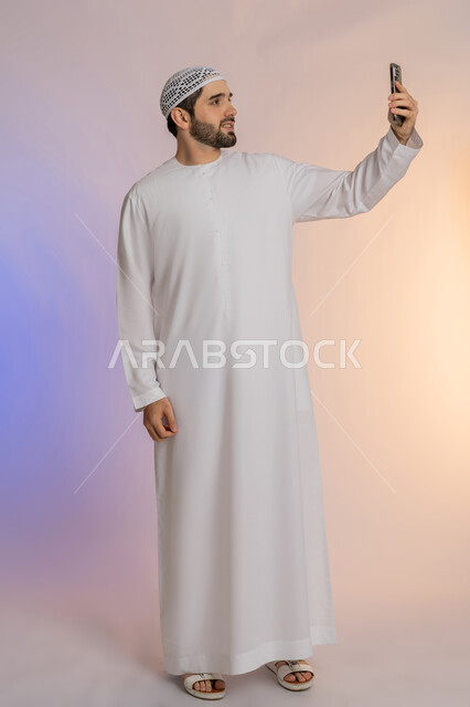 Taking a selfie using a mobile phone, making a video call and communicating with family and friends, integrating modern technology and techniques into daily life, portrait of an Arab Gulf Emirati man wearing a kandura and hat holding a mobile phone, full body, colorful background
