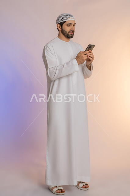 Using modern technologies and devices, integrating technology into daily life, browsing applications and social networking sites on the Internet, chatting, conversations and getting to know each other, portrait of an Arab Gulf Emirati man wearing a kandura and hat using a mobile phone, full-length body, colored background