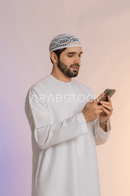 Browsing applications and social networking sites on the Internet, using modern technologies and devices, integrating technology into daily life, chatting, conversations and getting to know each other, side portrait of an Arab Gulf Emirati man wearing a kandura and hat using a mobile phone, colorful background