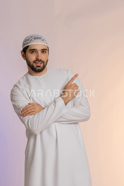 تحديد موقع شيء معين، إشارات وحركات يدين تدل على هذا المكان (هنا)، بورتريه لرجل عربي خليجي اماراتي يرتدي الكندورة والقبعة البيضاء يشير باصبع السبابة إلى الأعلى، مفهوم الأناقة والاهتمام بالمظهر الخارجي، خلفية ملونة