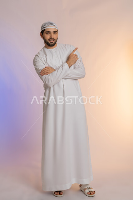 النظر الى الكاميرا بايماءات الثقة بالنفس، إشارات وحركات يدين تدل على هذا المكان (هنا)، تحديد موقع شيء معين، بورتريه لرجل عربي خليجي اماراتي يرتدي الكندورة والقبعة البيضاء يشير باصبع السبابة إلى الأعلى، الاهتمام بالمظهر الخارجي، الجسم بالطول الكامل، خلفية ملونة