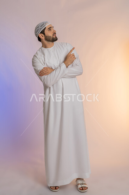 إشارات وحركات يدين تدل على هذا المكان (هنا)، تحديد موقع شيء معين، بورتريه لرجل عربي خليجي اماراتي يرتدي الكندورة والقبعة البيضاء يشير باصبع السبابة إلى الأعلى، مفهوم الأناقة والاهتمام بالمظهر الخارجي، الجسم بالطول الكامل، خلفية ملونة