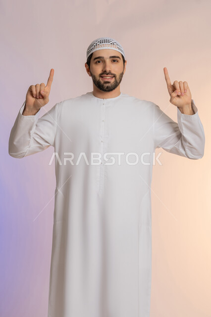 الأناقة والاهتمام بالمظهر الخارجي، حركات يدين واشارات تدل على هذا المكان (هنا)، تحديد موقع شيء معين، بورتريه لرجل عربي خليجي اماراتي يرتدي الكندورة والقبعة يشير باصبع السبابة إلى الأعلى، خلفية ملونة