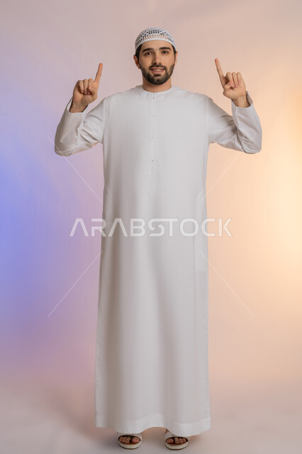 تحديد موقع شيء معين، حركات يدين واشارات تدل على هذا المكان (هنا)، بورتريه لرجل عربي خليجي اماراتي يرتدي الكندورة والقبعة يشير باصبع السبابة إلى الأعلى، مفهوم الأناقة والاهتمام بالمظهر الخارجي، الجسم بالطول الكامل، خلفيه ملونة