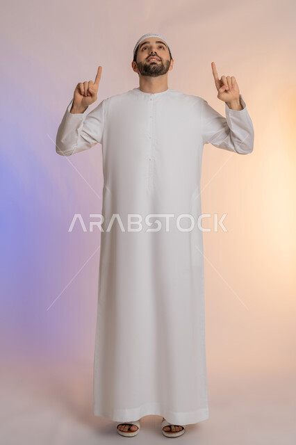 حركات يدين واشارات تدل على هذا المكان (هنا)، تحديد موقع شيء معين، بورتريه لرجل عربي خليجي اماراتي يرتدي الكندورة والقبعة يشير باصبع السبابة إلى الأعلى، مفهوم الأناقة والاهتمام بالمظهر الخارجي، الجسم بالطول الكامل، خلفيه ملونة