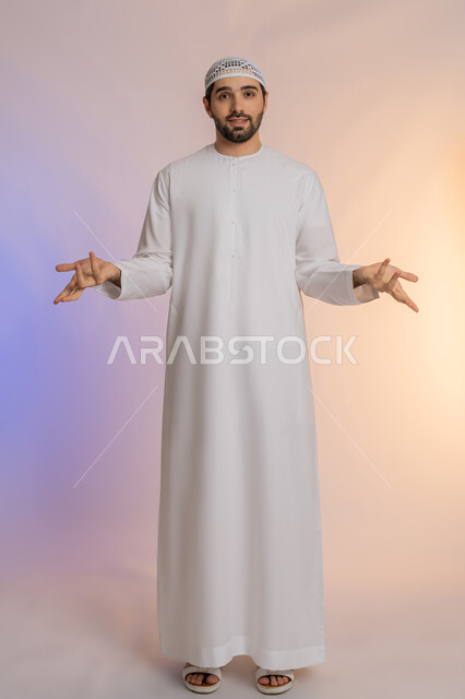 Standing with gestures of surprise and astonishment, feeling happy and pleased to hear the expected news, portrait of an Arab Gulf Emirati man wearing a kandura and hat, opening his hands with expressions of shock, astonishment and amazement, surprised and surprised by something, full body length, colored background