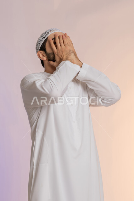 الشعور بالخجل والإحراج، إغماض العينين وعدم الرغبة بالرؤية، البكاء ومشاعر الحزن، التعرض لصدمة وعدم التصديق، بورتريه لرجل عربي خليجي اماراتي يرتدي الكندورة والقبعة البيضاء يغطي وجهه بيديه، خلفية ملونة
