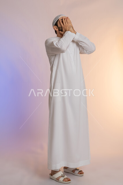 إغماض العينين وعدم الرغبة بالرؤية، الشعور بالخجل والإحراج، البكاء ومشاعر الحزن، التعرض لصدمة وعدم التصديق، بورتريه لرجل عربي خليجي اماراتي يرتدي الكندورة والقبعة البيضاء يغطي وجهه بيديه، الجسم بالطول الكامل، خلفية ملونة