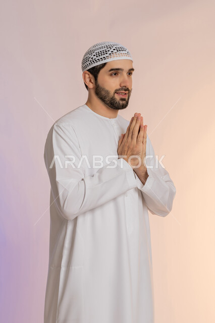 مفهوم الشكر والامتنان، الطلب بترجي وتذلل وخضوع، الوقوف بإيماءات الدعاء والتوسل والاسترحام، بورتريه لرجل عربي خليجي اماراتي يرتدي الكندورة والقبعة يرفع يديه بخشوع واستعطاف، طلب العون والرغبة في تحقيق الأمنيات، خلفية ملونة