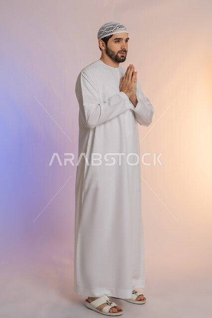 الطلب بترجي وتذلل وخضوع، مفهوم الشكر والامتنان، الوقوف بإيماءات الدعاء والتوسل والاسترحام، بورتريه لرجل عربي خليجي اماراتي يرتدي الكندورة والقبعة يرفع يديه بخشوع واستعطاف، طلب العون والرغبة في تحقيق الأمنيات، الجسم بالطول الكامل، خلفية ملونة