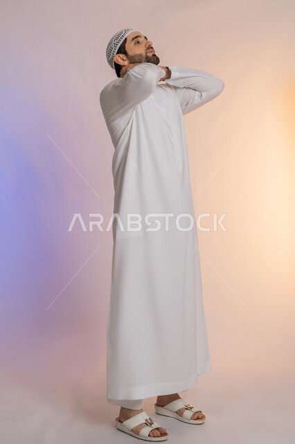 Bone and spine pain, arthritis and muscle inflammation in the neck, the need to go to the doctor and get examined, the need to relax and take a rest, a portrait of an Arab Gulf Emirati man wearing a kandura and a hat, putting his hand around his neck with gestures of pain, full-length bodycolored background