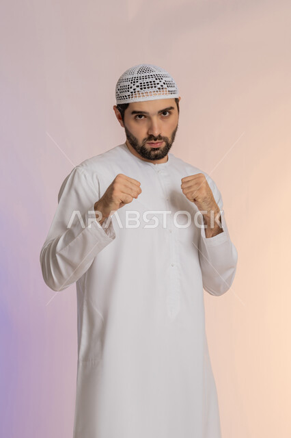 إغلاق اليدين والاستعداد للهجوم والمواجهة، احتراف رياضة الملاكمة، محاولة الدفاع عن النفس، مفهوم القوة والرجولة، بورتريه لرجل عربي خليجي اماراتي يرتدي الكندورة والقبعة ينظر للكاميرا بإيماءات الجدية والغضب،  خلفية ملونة