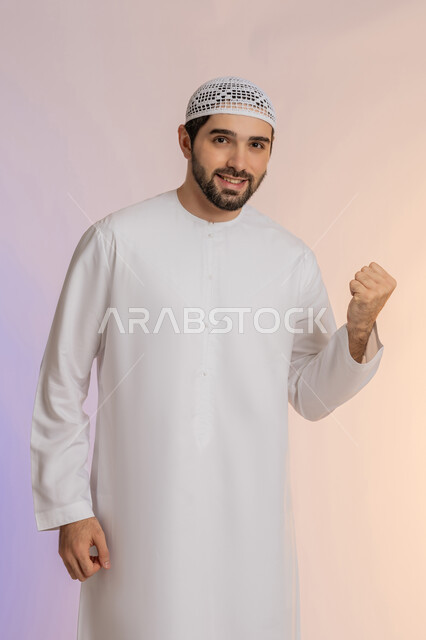 النجاح والفوز بشيء ما منتظر، الفرح الهستيري بتحقيق الأهداف والأحلام، الحماس والسعادة العارمة، بورتريه لرجل عربي خليجي اماراتي مبتسم يرتدي الكندورة والقبعة  يرفع قبضته في الهواء وينظر إلى الكاميرا بإيماءات السرور، خلفية ملونة