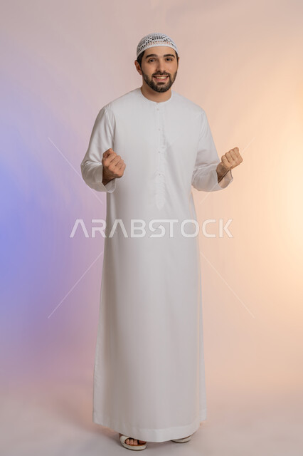 الحماس والسعادة العارمة، الفرح الهستيري بتحقيق الأهداف والأحلام، النجاح والفوز بشيء ما منتظر، بورتريه لرجل عربي خليجي اماراتي مبتسم يرتدي الكندورة والقبعة  يرفع قبضتيه في الهواء وينظر إلى الكاميرا بإيماءات السرور، الجسم بالطول الكامل، خلفية ملونة