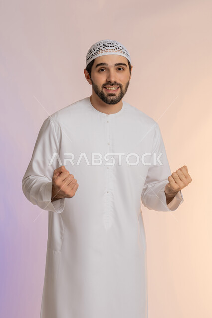 Hysterical joy of achieving goals and dreams, enthusiasm and overwhelming happiness, success and winning something is waiting, portrait of a smiling Arab Gulf Emirati man wearing a kandura and a hat raising his fists in the air and looking at the camera with gestures of pleasure, colorful background