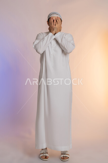 إغماض العينين وعدم الرغبة بالرؤية، الشعور بالخجل والإحراج، البكاء ومشاعر الحزن، التعرض لصدمة وعدم التصديق، بورتريه لرجل عربي خليجي اماراتي يرتدي الكندورة و القبعة يغطي وجهه بيديه، الجسم بالطول الكامل، خلفية ملونة